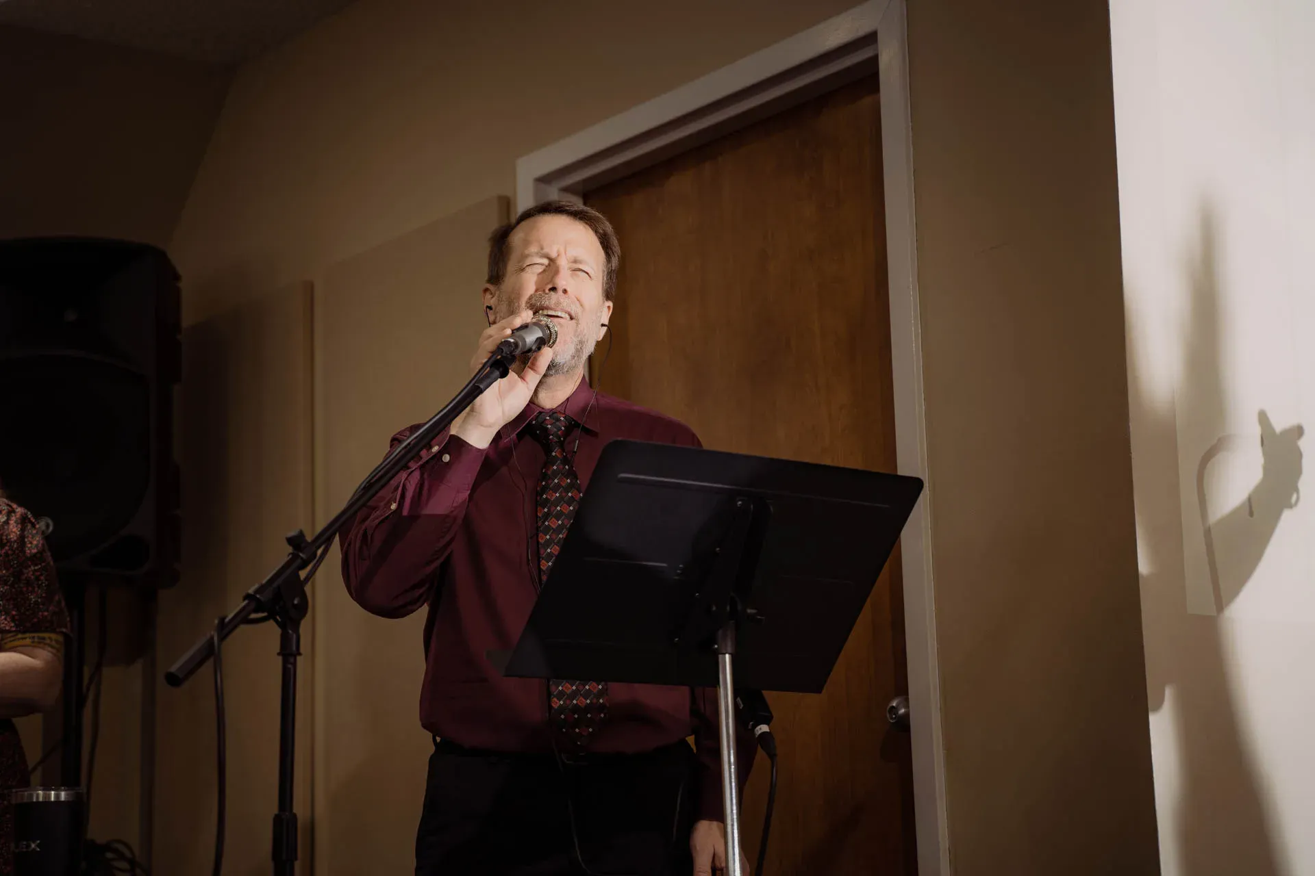 A man in a burgundy dress shirt and patterned tie sings into a microphone on a boom stand, eyes closed and head tilted up, behind a black music stand.